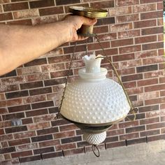 a hand holding a white light fixture in front of a brick wall with a chain hanging from it