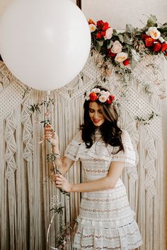 a woman in a white dress is holding a balloon and standing next to a wall with flowers on it