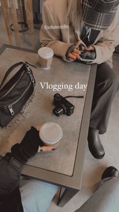 a woman sitting at a table with a camera and cell phone in her hand while another person holds a coffee cup