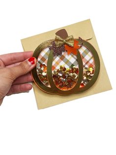 a hand holding an orange and gold pumpkin shaped card with sequins on it