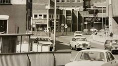 black and white photograph of cars driving down the street in front of buildings on both sides