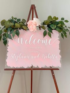 a welcome sign with flowers and greenery hanging from it's sides on a easel