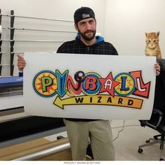 a man holding up a sign that says pinball wizard with a cat in the background