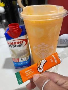 a hand holding an orange drink next to a carton of cremel protein