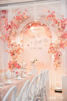 a table set up with white chairs and pink flowers on the wall behind it is an archway