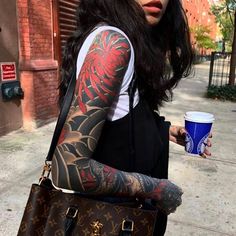 a woman with tattoos on her arm holding a coffee cup and handbag while walking down the street