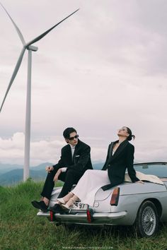 two people sitting on top of a car in front of a wind turbine