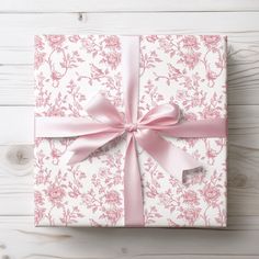 a gift wrapped in pink and white floral paper with a bow on the top, sitting on a wooden surface