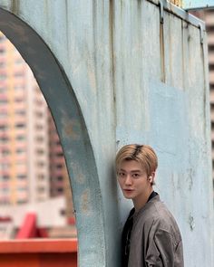 a young man leaning against the side of a blue metal structure with buildings in the background