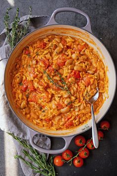 a pot filled with pasta and tomatoes on top of a table next to some herbs