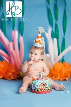 a baby sitting on the ground wearing a birthday hat and eating cake with balloons in the background