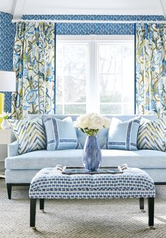 a blue and white living room with floral curtains on the window sill, a large flower vase in front of the couch