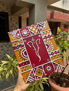 a hand holding up a decorative tile in front of a potted plant