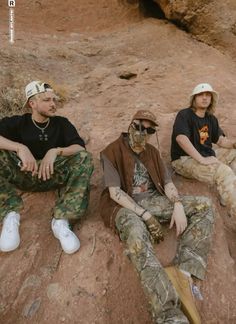 three men sitting on the ground with their feet up and one man wearing camouflage clothing