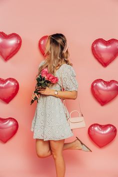 a woman in a white dress is holding flowers and standing against a pink background with heart shaped balloons