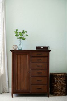 a wooden cabinet sitting in front of a window next to a basket with a plant