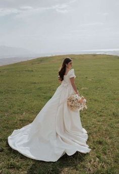 a woman in a wedding dress standing on top of a grass covered field holding a bouquet