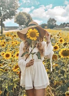 a woman standing in a sunflower field with her hat on and holding a flower