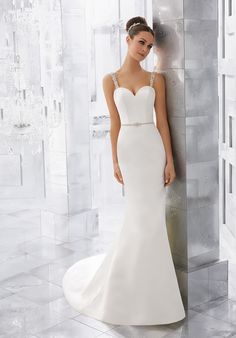 a woman standing next to a wall wearing a white wedding dress with straps and beading