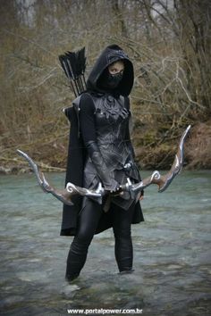 a woman dressed in black and holding two arrows, standing in the water with trees behind her