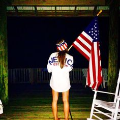 a woman holding an american flag and standing next to a white rocking chair in the dark