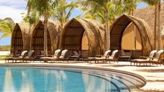 an outdoor swimming pool with lounge chairs and palm trees surrounding the poolside, surrounded by thatched huts