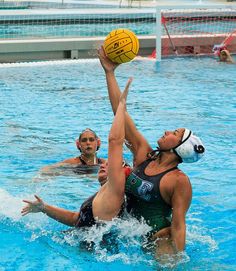 two women playing water polo in a pool with one trying to block the ball from her