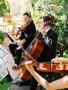 two men in suits playing violin and another man wearing a tuxedo sitting on a bench