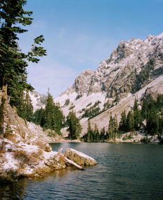 a mountain lake surrounded by snow covered mountains and pine trees in the foreground, with a rock outcropping on the right side