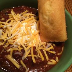 a green bowl filled with chili and cheese next to a baguette on top of a wooden table