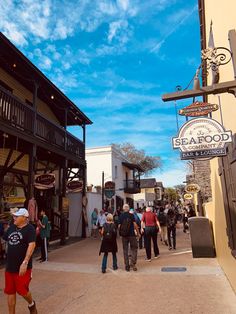 many people are walking down the street in front of some shops and restaurants on a sunny day