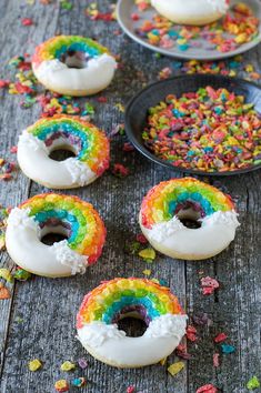 rainbow sprinkle covered donuts sitting on top of a wooden table next to bowls of sprinkles