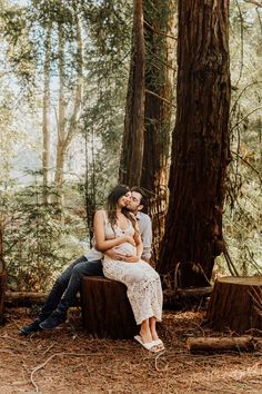 a man and woman are sitting on a log in the woods with trees behind them