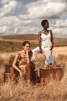 two women sitting on suitcases in the middle of a field with clouds above them