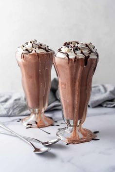 two glasses filled with chocolate milkshakes on top of a white table next to silverware