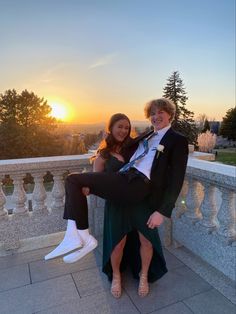 a man and woman posing for a photo on a balcony with the sun setting in the background