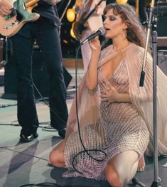 a woman sitting on the ground while singing into a microphone and holding a guitar in her hand