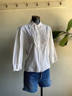 "Vintage 1970s Unbranded White Cotton Ruffle Blouse. White in color made from cotton fibers. Brand is unknown (tag has been cut). Long sleeve blouse with ruffled accents along front. Button down closure along front and cuffs. Overall condition of garment is good with only minor flaws that include; blouse is missing one of the buttons on the right cuff as shown in photos. No rips, tears, or staining. Please see all photos for details. Refer to measurements below to ensure a proper fit (modeled on Classic Cotton Top With Ruffled Collar, Classic Cotton Shirt With Ruffles, Cotton Button-up Tops With Lace Collar, Victorian Cotton Tops For Spring, Spring Victorian Cotton Tops, Cotton Blouse With Ruffled Collar, Cotton Blouse With Ruffled Collar And Details, Cotton Ruffle Blouse For Daywear, Retro Cotton Top With Lace Collar