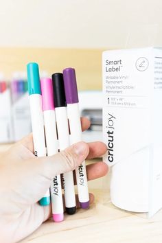 a person holding four different colored pens in front of a white box on a table