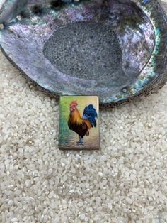 a small square brooch with a rooster on it's side sitting on some white rice