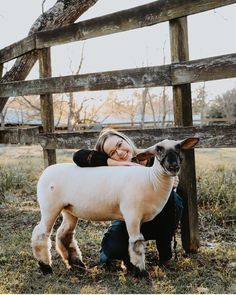 a woman holding a sheep in front of a wooden fence with grass on the ground