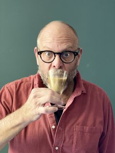 a man with glasses and a beard is drinking something from a glass in front of his face