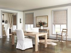 a dining room with white chairs and wooden table surrounded by windows covered in roman shades