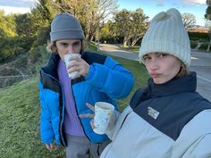 two people standing in the grass drinking from coffee mugs and looking at the camera