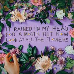 someone is writing on a sign surrounded by flowers and leaves that says it rained in my head for a month but now i look at all the flowers