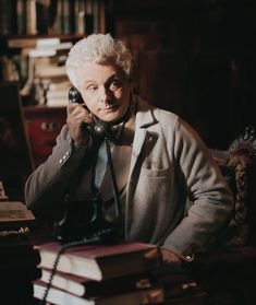 an old man sitting at a desk talking on the phone and holding a stack of books