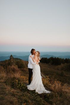 a bride and groom standing on top of a hill in the mountains at sunset with their arms around each other