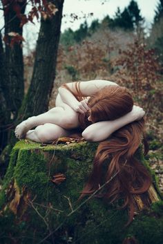 a woman with red hair laying on top of a moss covered stump in the woods