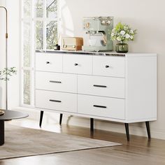 a white dresser sitting in a living room next to a table with flowers on it