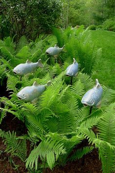 several metal fish sitting on top of ferns in a garden with trees and bushes behind them